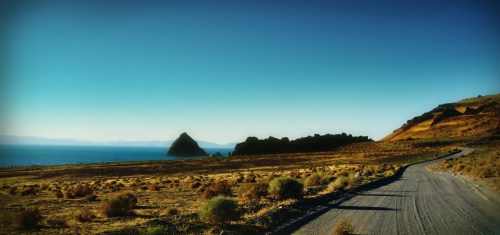 A scenic view of a gravel road leading to a lake, with mountains and a clear blue sky in the background.
