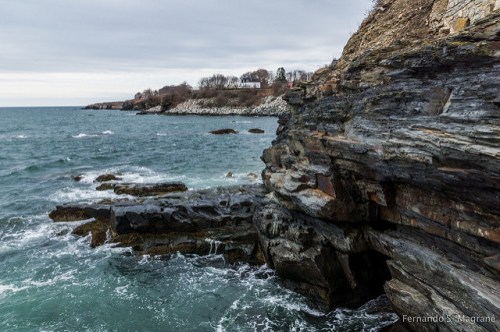 Rocky coastline with waves crashing against the shore under a cloudy sky, featuring distant houses on the cliffs.