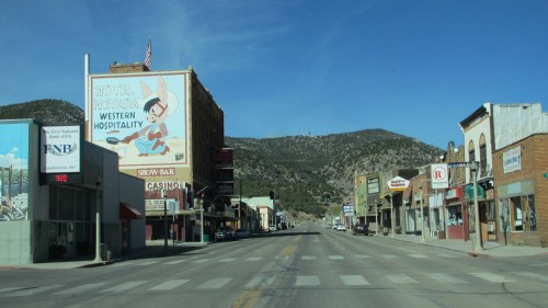A quiet street scene featuring shops, a casino, and a large billboard with a cartoon character in a mountainous area.