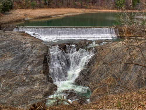 A serene waterfall cascading over rocks into a calm, greenish pond surrounded by bare trees and grass.