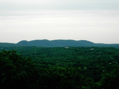 A panoramic view of rolling green hills under a cloudy sky, with distant mountains on the horizon.