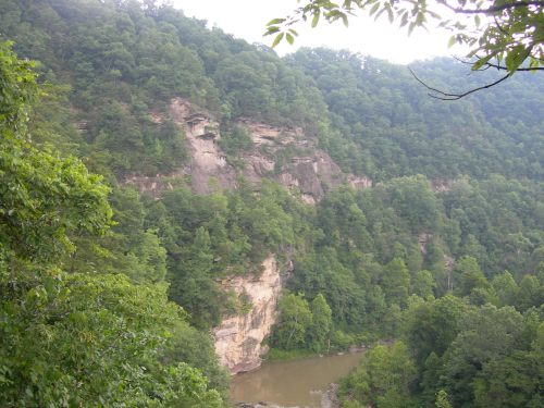 Lush green forested hills surround a winding river, with rocky cliffs visible in the background under a cloudy sky.