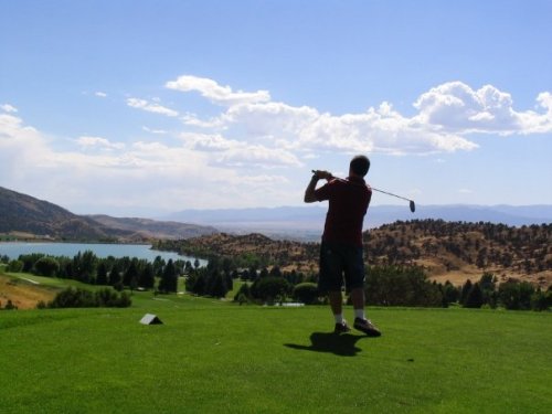 A golfer swings a club on a lush green course with mountains and a lake in the background under a blue sky.