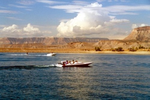 Two boats speed across a calm lake, surrounded by rocky cliffs and fluffy clouds in a blue sky.