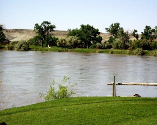 A calm river flows through a green landscape, surrounded by trees and distant hills under a clear sky.