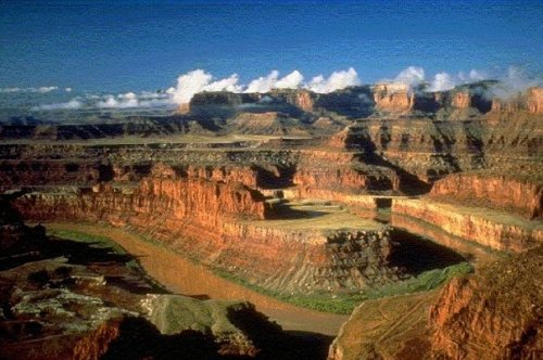 A panoramic view of a canyon with red rock formations and a winding river under a blue sky with clouds.