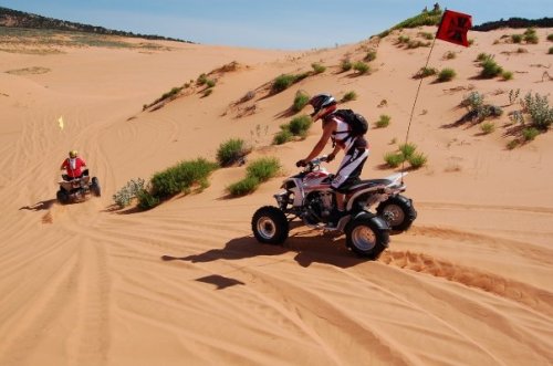 Two riders on ATVs navigate sandy dunes under a clear blue sky, with vegetation scattered around.