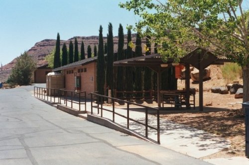 A pathway leads to a shaded picnic area with tall trees and a building against a rocky landscape.