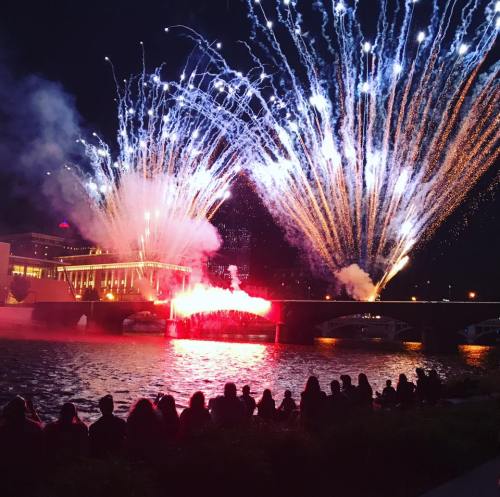 Colorful fireworks illuminate the night sky over a river, with spectators sitting along the water's edge.