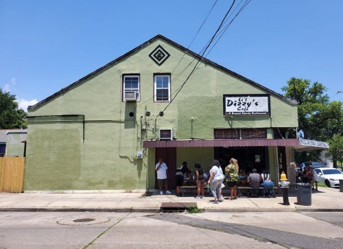 A green building with a sign reading "Lil Dizzy's Cafe," featuring people sitting outside on a sunny day.