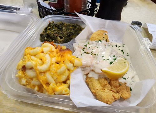 A takeout container with fried fish topped with sauce, macaroni and cheese, and collard greens, garnished with a lemon slice.