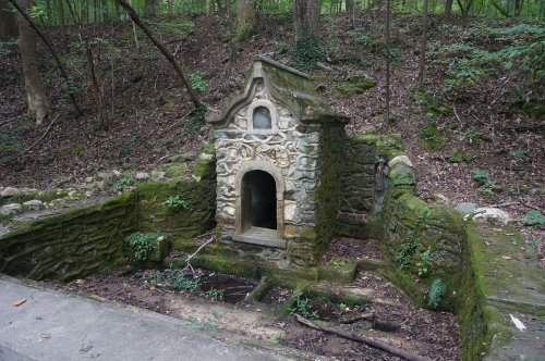 A moss-covered stone structure in a wooded area, partially overgrown with plants and surrounded by trees.