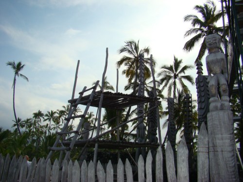A wooden structure with a ladder surrounded by palm trees and carved wooden figures, under a cloudy sky.