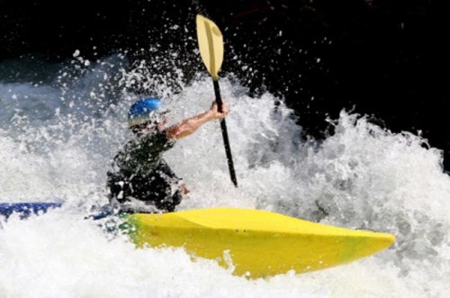 A person in a blue helmet paddles a yellow kayak through turbulent white water rapids.