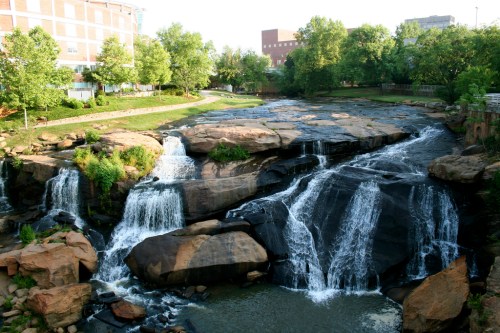 A serene waterfall cascades over rocky terrain, surrounded by lush greenery and urban buildings in the background.