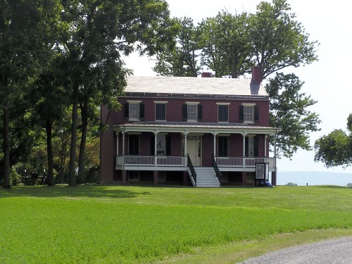 A two-story red brick house with a white porch, surrounded by green grass and trees on a sunny day.