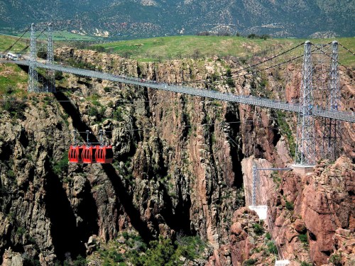 A cable car crosses a deep canyon, with rocky cliffs and green hills in the background under a clear blue sky.