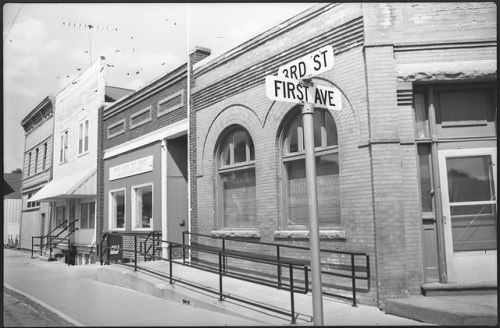 Black and white photo of a street corner with buildings and a sign for 3rd St and First Ave.