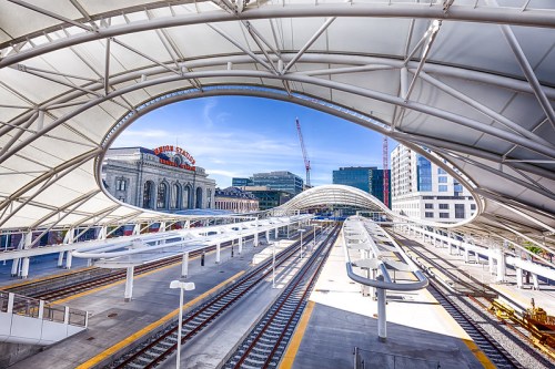 A modern train station with curved roofs, tracks, and a historic building in the background under a clear blue sky.