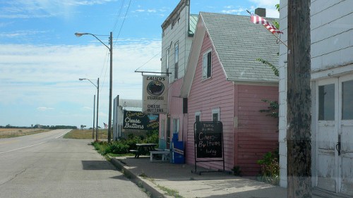 A small roadside shop with a pink building, signs for cheese and butter, and a flag waving in the breeze.