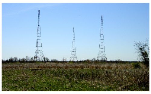 Three tall radio towers stand in a grassy field under a clear blue sky.