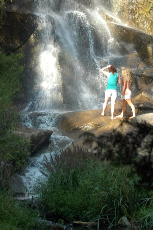 Two women stand near a waterfall, surrounded by lush greenery and rocky terrain. Sunlight glistens on the water.