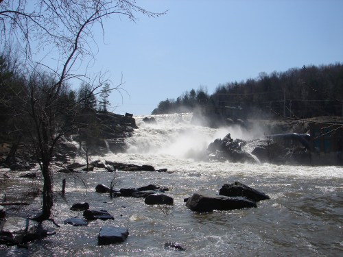 A powerful waterfall cascades over rocks, surrounded by trees and mist under a clear blue sky.