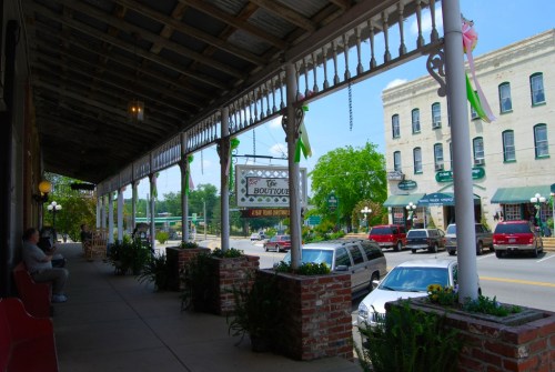 A charming street view with a covered porch, plants, and shops in a small town on a sunny day.