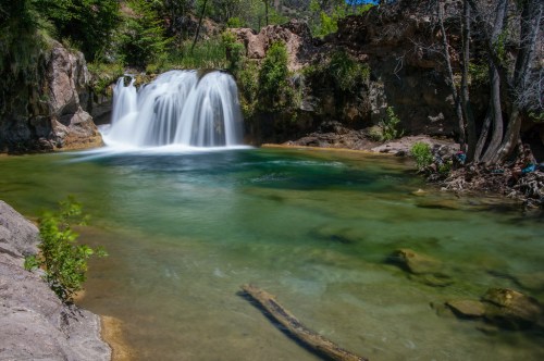 A serene waterfall cascades into a clear pool, surrounded by lush greenery and rocky terrain under a bright blue sky.