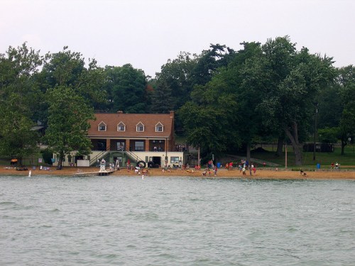 A lakeside scene featuring a beach, a building, and people enjoying the outdoors among trees and water.