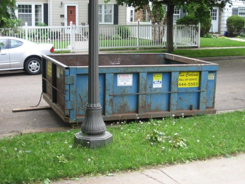 A large blue dumpster on a residential street, with a lamppost nearby and grass scattered around.