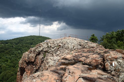 A rocky cliff with a metal railing overlooks a lush green landscape under dark, stormy clouds.