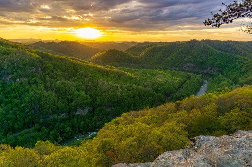 A scenic view of lush green mountains and a winding river at sunset, with a dramatic sky overhead.