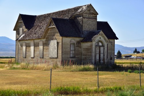 Abandoned wooden building with boarded windows, surrounded by tall grass and a fence, set against a clear blue sky.