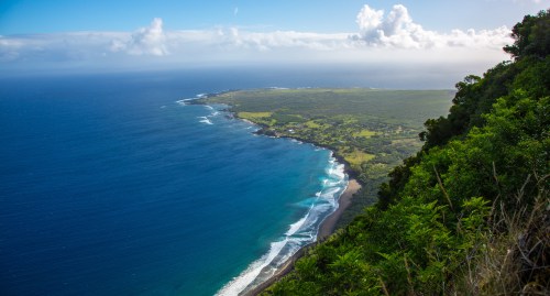 Aerial view of a lush coastline with waves crashing on the shore, surrounded by green hills and a blue sky.