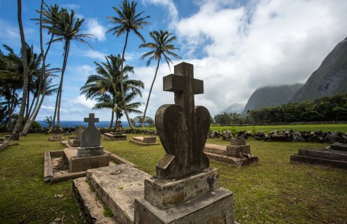 Graveyard with stone crosses, surrounded by palm trees and mountains under a cloudy sky.
