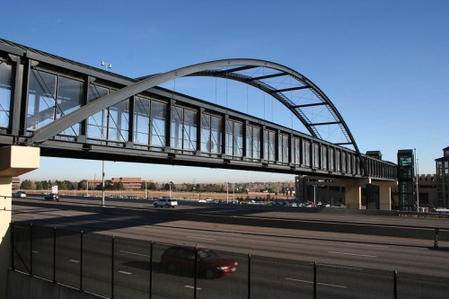 A modern pedestrian bridge with an arched design spans over a busy highway, connecting buildings on either side.