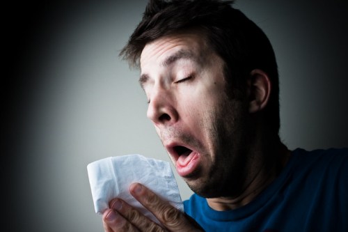 A man with a pained expression sneezes into a tissue, eyes closed, against a dark background.