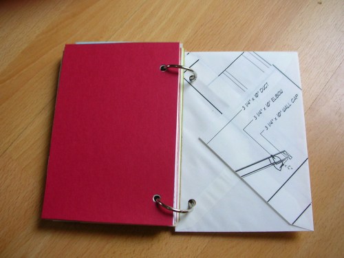A partially open binder with a red cover and a sketch on white paper inside.