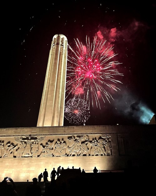 Fireworks illuminate the night sky above a tall monument, with silhouettes of people in the foreground.