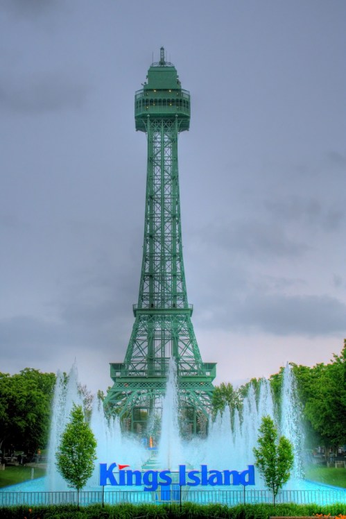 Eiffel Tower replica at Kings Island amusement park, surrounded by fountains and greenery under a cloudy sky.
