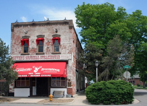 A historic brick building with red awnings, featuring a steakhouse sign and surrounded by trees and greenery.