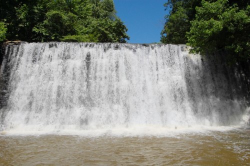 A cascading waterfall flows over a rocky ledge into a brown river, surrounded by lush green trees under a clear blue sky.