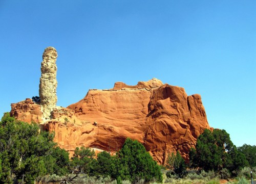 A tall rock formation stands beside a large, reddish cliff under a clear blue sky, surrounded by green shrubs.