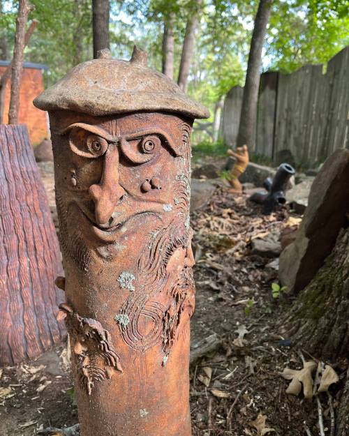 A whimsical, carved wooden figure with a face, surrounded by trees and rustic sculptures in a natural setting.