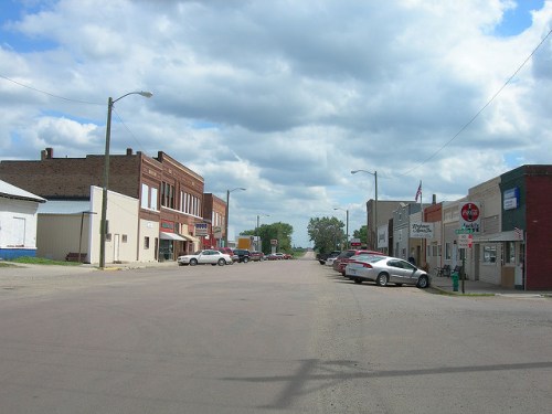 A quiet small-town street lined with shops and parked cars under a cloudy sky.