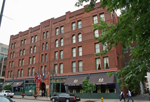 Historic red brick hotel with flags, awnings, and greenery in front, located in an urban setting.