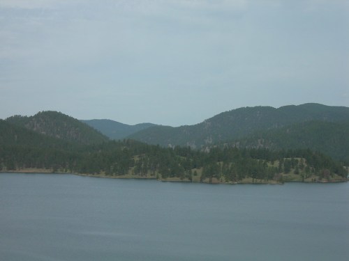 A serene landscape featuring a calm lake surrounded by green hills and distant mountains under a cloudy sky.