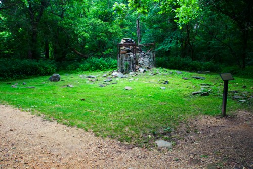 A grassy area with scattered rocks and a metal structure surrounded by trees, with a sign nearby.