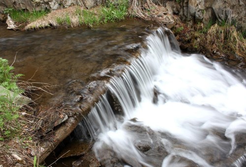 A serene stream flows over rocks, creating a gentle waterfall surrounded by greenery and rugged terrain.